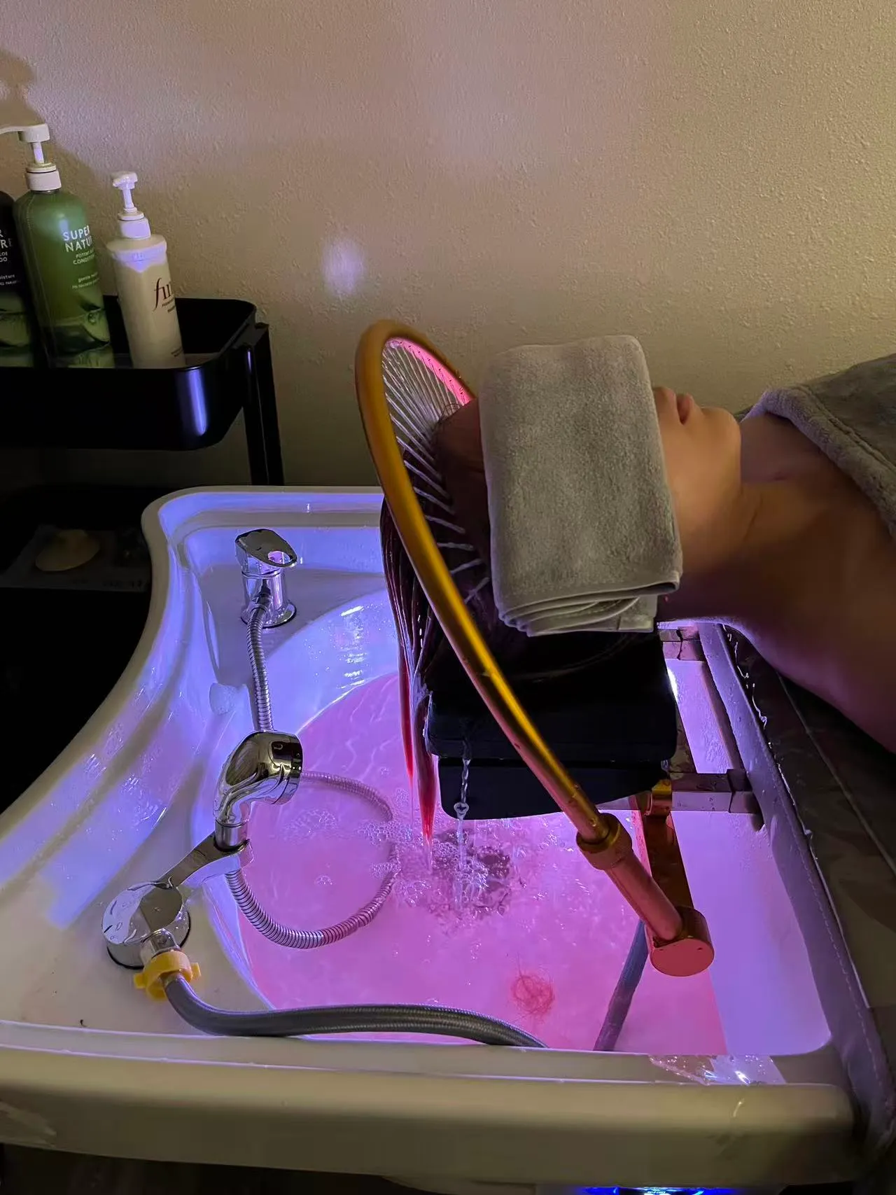Water from the gold waterfall arch running through a client's hair into a rose-lit head spa basin at O2 Head & Beauty Spa in Arcadia, a towel folded over the eyes