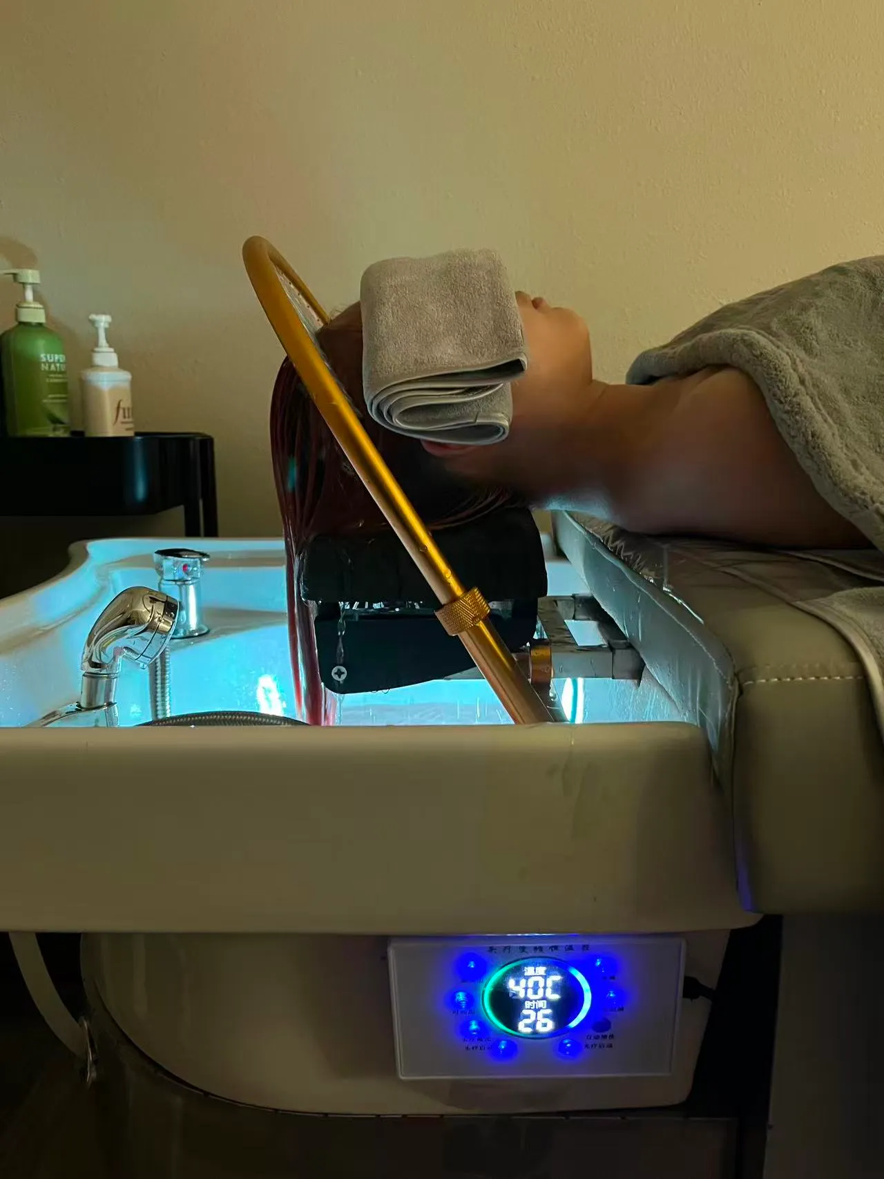 Client resting at an O2 head spa basin with a folded towel over the eyes while the blue panel shows the water temperature